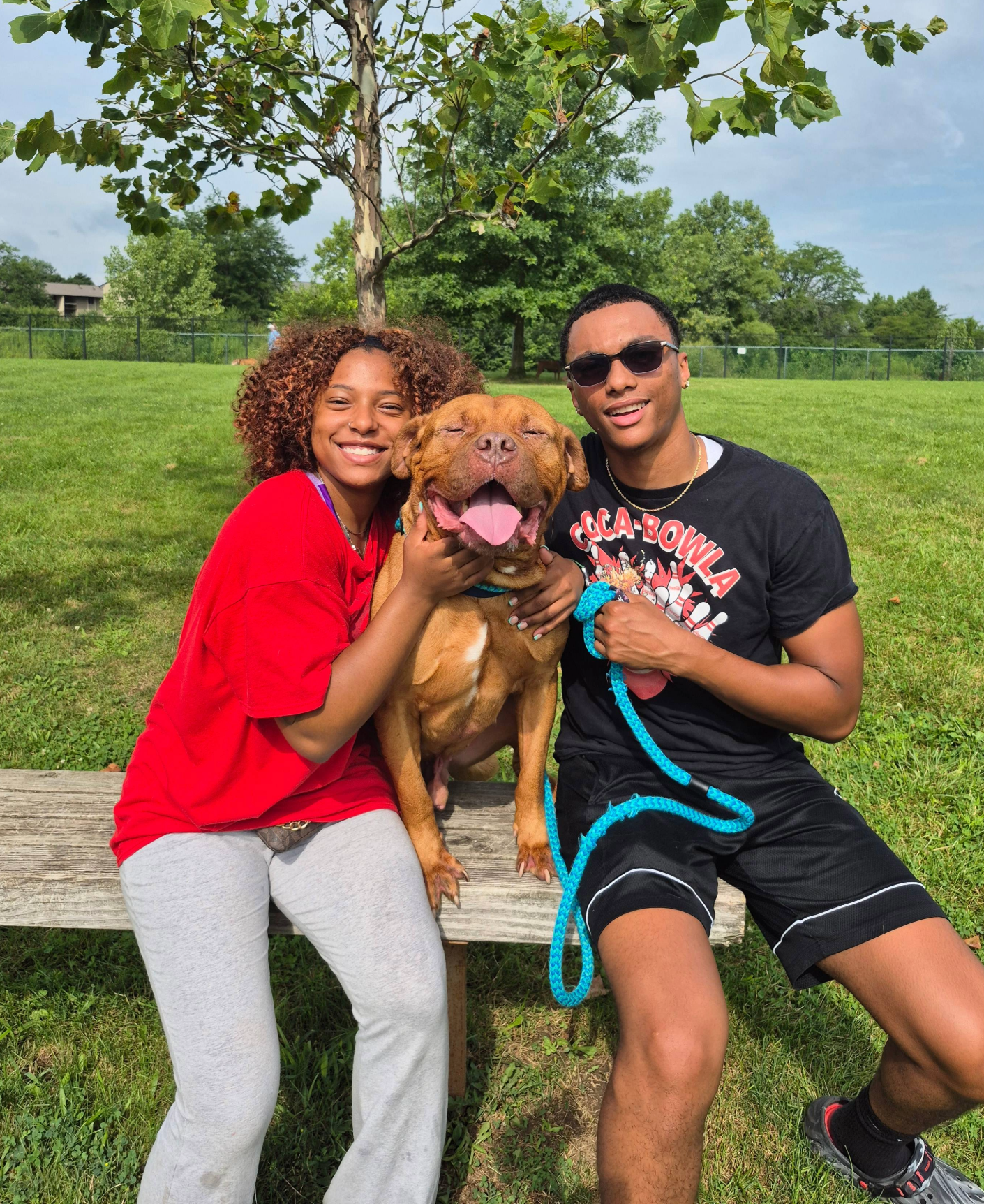 Photo of a man and a woman sitting with a brown dog outside on a bench smiling
