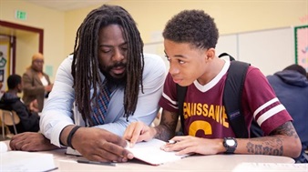 A man helps a teenage boy with his homework at a table