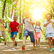 A group of children with smiling faces run in front of trees and a shining sun. 